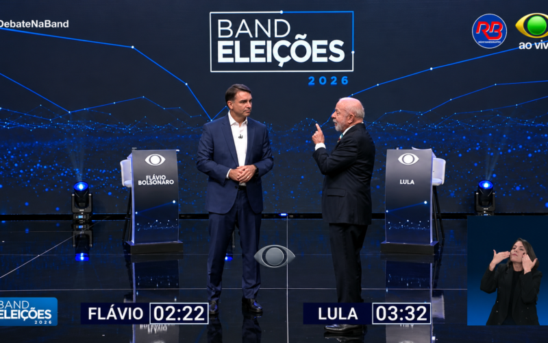 Lula e Flávio Bolsonaro de pé no cenário do debate da Band, com os cronômetros de fala na tela