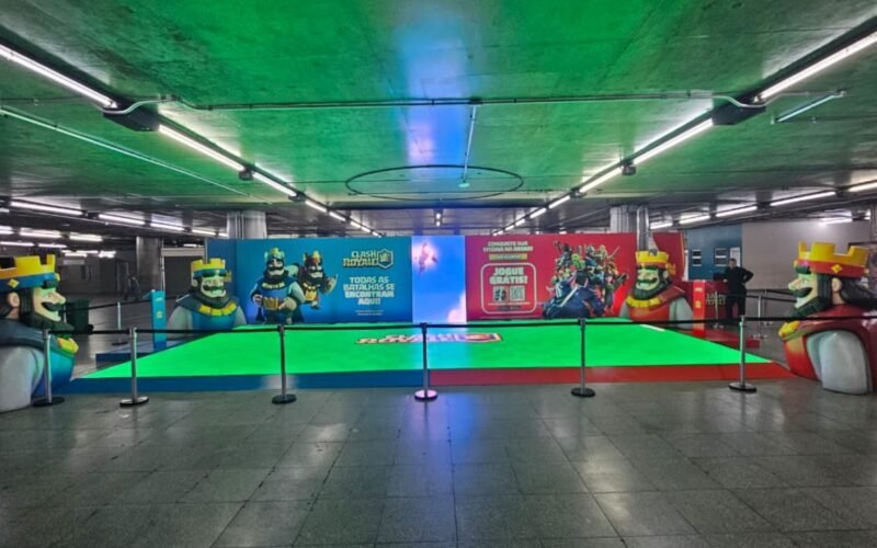 Arena de piso verde iluminado montada em estação de metrô, com cenografia colorida de jogo ao fundo
