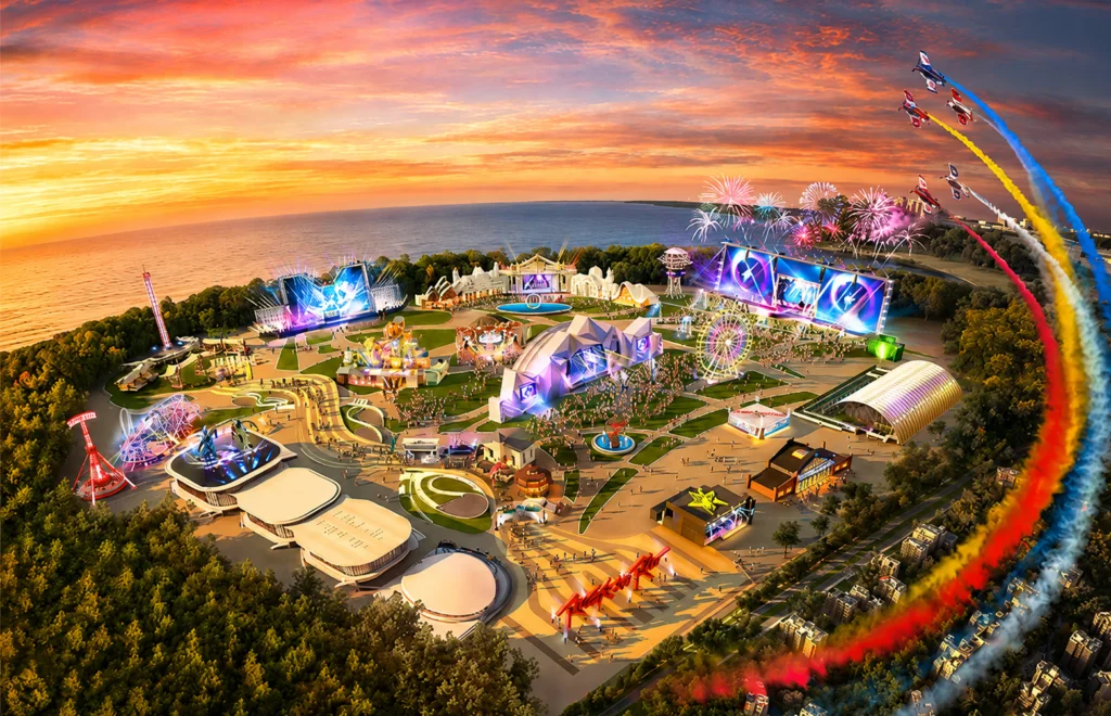Vista aérea de um festival de música à beira-mar, com palcos iluminados, roda-gigante e rastros coloridos de aviões ao pôr do sol