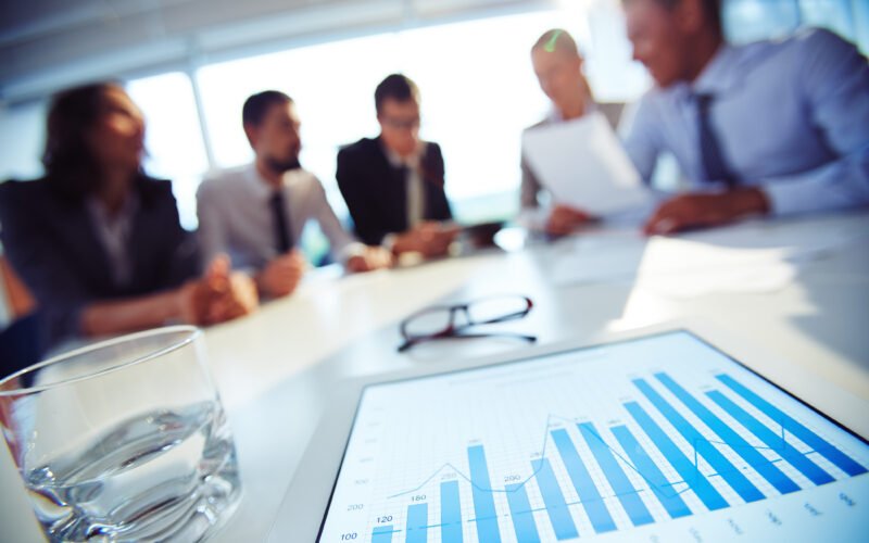 Pessoas em reunião ao redor de uma mesa com gráfico de barras impresso, sobre o planejamento de mídia de 2027